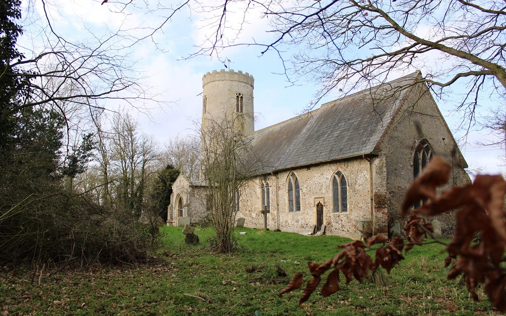 Ilketshall Saint Margaret Church and churchyard with ancient gravestones. The church is built of stone and cobble. It has a round tower and prominent porch. In the foreground is a branch of tan-coloured beech leaves and some evergreen shrubs. In the background are mature winter deciduous trees.