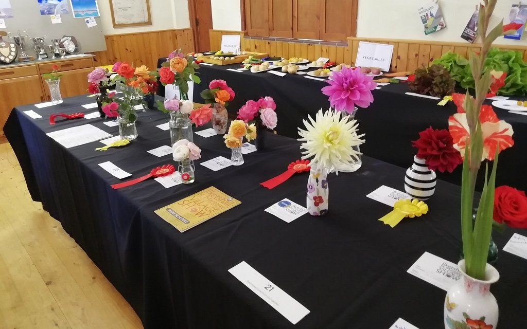 Colourful flower displays in vases on a long table in a wood-panelled room. The displays are accompanied by information cards and some have red and yellow rosettes next to them. In the background is another table with displays of vegetables. The tables are draped with black cloths. some silver cups and shields are visible to the left in the background.