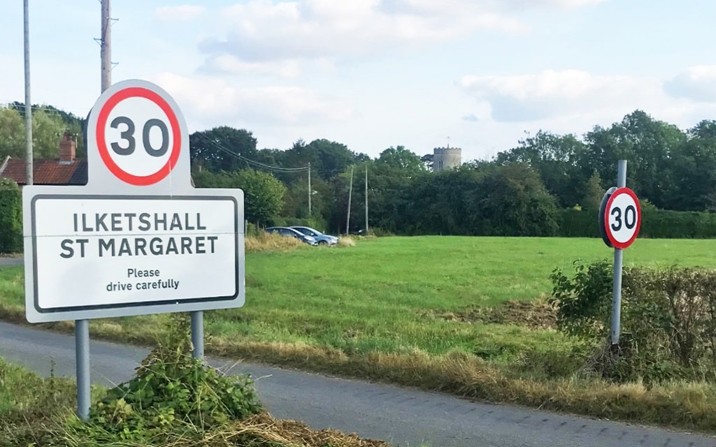 Entry to the village maked with a road sign displaying the name 'Ilketshall St Margaret' and a 30mph speed limit sign. The surrounding scenery comprises a green arable field and mature deciduous trees in full leaf. The top of the church tower can be seen in the background.