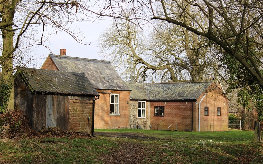 A single-storey building of red brick, with extensions of various heights, angles and ages. It is partially obscured by a small shed in the foreground. There is a mature deciduous tree behind the building. The building is lit by winter sunshine.